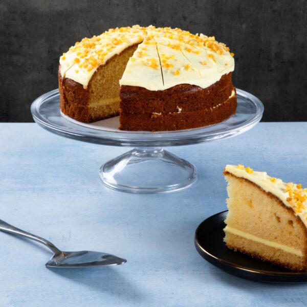 Orange drizzle cake with creamy frosting and orange zest on a glass cake stand.