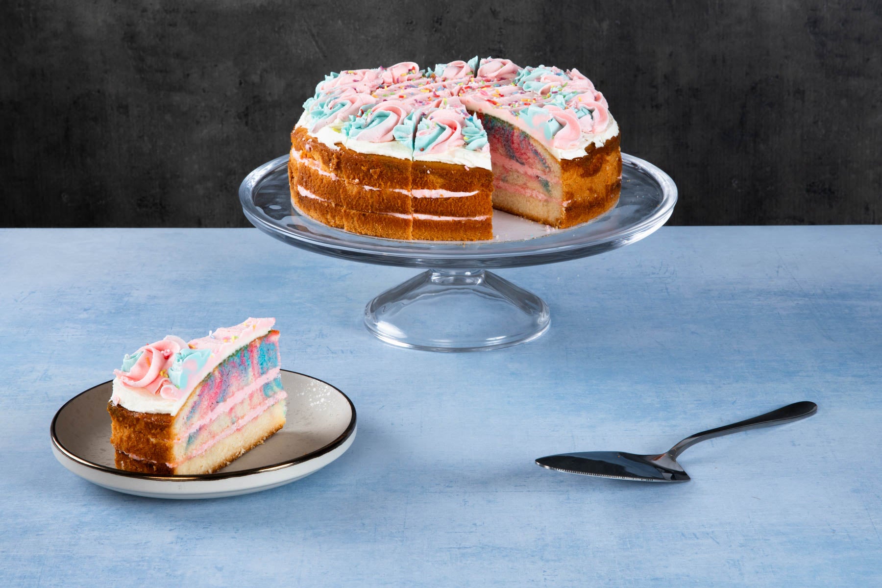 LHMC008-150.jpg Rainbow cake with colourful layers and white frosting on a glass cake stand.