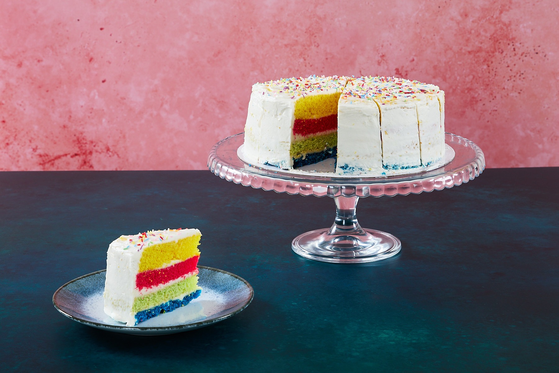 Rainbow Cake Rainbow cake with colourful sponge layers and sprinkles on a glass cake stand.